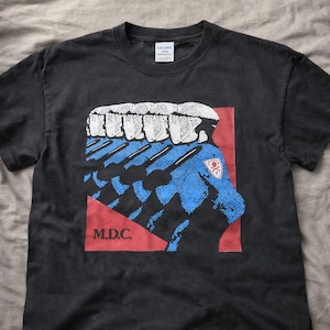 May include: Black t-shirt featuring a graphic design with blue and white elements on a red background. The design includes the text "M.D.C." and a badge. The shirt is made of a soft material.