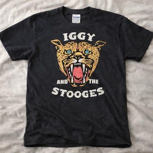 Puede incluir: Camiseta negra con un gráfico de leopardo rugiente con ojos azules y el texto "IGGY AND THE STOOGES" en blanco. La camiseta es de cuello redondo y parece ser de algodón.
