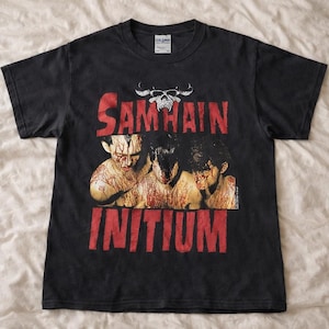 Puede incluir: Camiseta negra Gildan con el nombre de la banda "Samhain" en letras rojas en bloque sobre un gráfico de tres figuras cubiertas de sangre. La palabra "INITIUM" está impresa en letras rojas en bloque debajo.