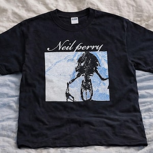 Puede incluir: Camiseta negra con el nombre "Neil perry" en escritura blanca sobre un gráfico. El gráfico es una ilustración en negro y azul de una figura sosteniendo un objeto. La camiseta está sobre una tela de color claro.