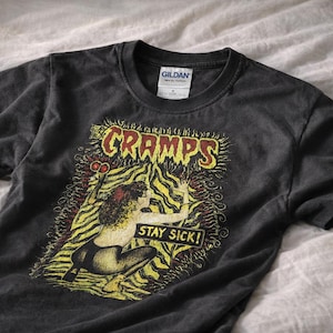 May include: Dark gray t-shirt featuring a graphic design with the word "CRAMPS" in red and yellow, and the words "STAY SICK!". The design includes a woman with scissors and a yellow and black background.