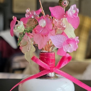 Puede incluir: Un arreglo floral decorativo con flores, hojas y cuentas de vidrio rosa y transparente. Las flores están dispuestas en un pequeño jarrón blanco con una cinta rosa atada alrededor de la base. El arreglo mide unos 13 cm de alto.
