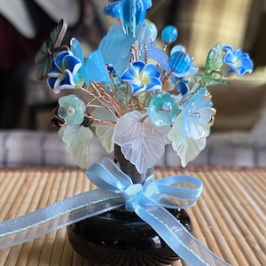 Könnte beinhalten: Ein dekoratives Glasblumenarrangement in einer kleinen schwarzen Vase, gebunden mit einem hellblauen Band. Das Arrangement zeigt blaue und weiße Blumen, hellblaue Blätter und Kupferdrahtstiele. Die Vase steht auf einer Bambusmatte.