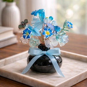 May include: A decorative floral arrangement in a small black vase, featuring blue and white glass flowers and leaves. The vase is tied with a light blue ribbon and sits on a marble tray. The arrangement includes various flower types and leaf shapes.