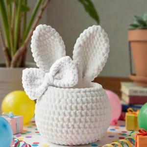 Peut inclure: Un panier lapin blanc crocheté avec des oreilles droites et un nœud. Le panier est rond et texturé, parfait pour la décoration de Pâques ou de printemps. Des ballons colorés et des confettis entourent le panier.