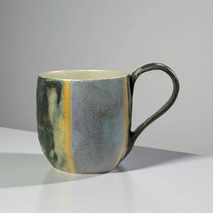 May include: A ceramic mug with a unique, textured finish. The mug features a colour palette of blue, green, and yellow vertical stripes. The handle is a dark, curved shape. The interior is a light cream colour. This handmade mug is perfect for coffee or tea.