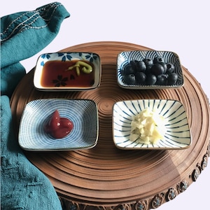 May include: Four small, square ceramic dishes with blue and white patterns, each containing different foods. One holds blueberries, another sauce with wasabi, a third ketchup, and the last, chopped garlic. The dishes sit on a wooden serving tray.