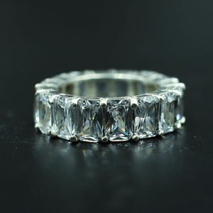 Könnte beinhalten: Ein silberfarbener Eternity-Ring mit einer durchgehenden Reihe klarer, rechteckiger Steine im Smaragdschliff, die in einer Krappenfassung sitzen. Der Ring hat eine polierte metallische Oberfläche und zeigt facettierte Steine, die das Licht reflektieren.