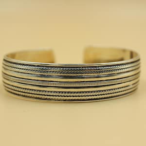 Könnte beinhalten: Silberfarbenes Armreif-Armband mit aufwendigen, geätzten Details. Das Armband hat mehrere Bänder mit strukturierten Mustern, darunter Linien und seilartige Designs. Das offene Design ermöglicht ein einfaches Tragen.