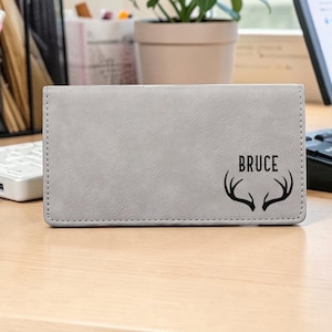 May include: A rectangular gray wallet featuring a smooth, matte finish with visible stitching along the edges. The front is engraved with the name BRUCE in a bold, sans-serif font positioned above a black graphic illustration of deer antlers.