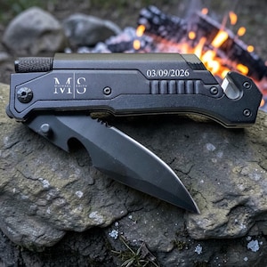 May include: A black multi-tool knife with a partially open blade rests on a gray rock. The tool has the initials "MS" and the date "03/09/2026" engraved on it. A campfire is visible in the background.