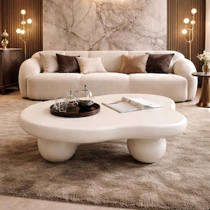 May include: A modern living room featuring a cream-colored, cloud-shaped coffee table with rounded legs. A cream-colored sofa with several throw pillows sits in the background. Decorative lamps and a rug complete the scene.