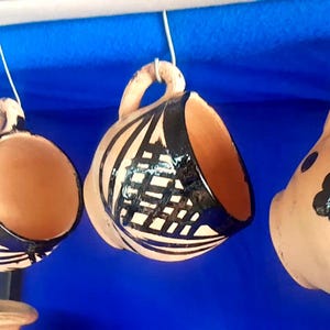 Puede incluir: Cuatro tazas de cerámica hechas a mano con asas, suspendidas por cuerdas. Las tazas tienen un color arcilla natural con diseños pintados en negro, incluyendo patrones geométricos y motivos florales. Los interiores son de un tono cálido y terroso.