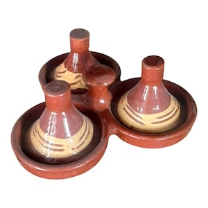 Può includere: Un server per condimenti in ceramica color terracotta con tre sezioni. Ogni sezione contiene un piccolo contenitore bulboso con un design a strisce color crema e bordeaux. Il server ha una maniglia centrale.