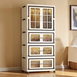 May include: A tall, off-white storage cabinet with multiple compartments. The cabinet has a dark brown trim and clear glass doors. The top section displays hanging clothes, while the lower sections contain various items, including stuffed animals and folded linens.