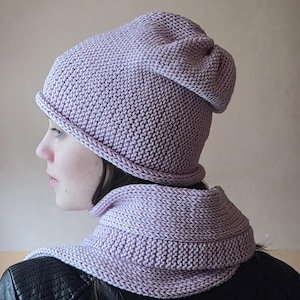 May include: A light purple knitted hat and cowl set. The hat is a beanie style with a folded brim and a slightly slouchy fit. The cowl is a neck warmer. Both pieces are made of a soft, textured yarn, perfect for cold weather.