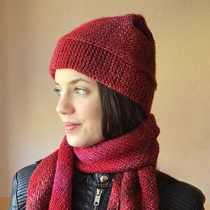 May include: A knitted red hat and scarf set. The hat is a close-fitting beanie style, and the scarf is long and rectangular. The set is made of a textured yarn with variations of red and pink hues. The person is wearing a black leather jacket.