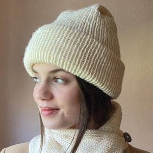 May include: Cream-colored knit beanie and matching scarf. The beanie has a folded brim and a ribbed texture. The scarf is wrapped around the neck and fastened with a wooden button. The set is made of a soft, warm material.