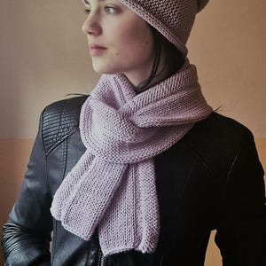 Puede incluir: Un conjunto de bufanda y gorro de punto de color lila. La bufanda está envuelta alrededor del cuello y el gorro está en la cabeza. El conjunto está hecho de un hilo suave y texturizado. La persona lleva una chaqueta de cuero negra.