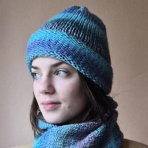 May include: A knitted hat and scarf set in shades of blue and purple. The hat is a beanie style with a folded brim. The scarf is a cowl neck design. The set is made of a soft, textured yarn, perfect for cold weather.