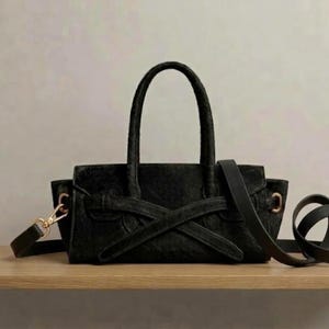 May include: A black handbag with a textured surface, featuring two top handles and a decorative crossover strap detail on the front. The bag includes a detachable shoulder strap with gold-toned metal hardware clips on the sides.