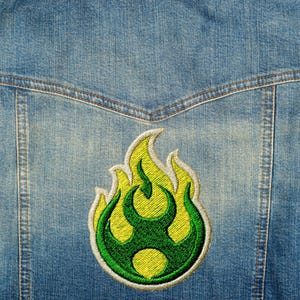 May include: Close-up of a denim jacket with a vibrant embroidered patch. The patch features a stylized flame design in shades of green and yellow, outlined in white. The jacket's blue denim fabric provides a contrasting backdrop.
