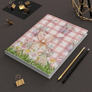 Op de afbeelding: Een hardcover notitieboek met een roze en wit geruit patroon en illustraties van een konijn, bloemen en strikken. Het notitieboek is omgeven door gouden en zwarte kantoorbenodigdheden, waaronder paperclips en potloden.