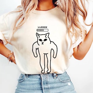 May include: Cream-colored t-shirt featuring a black cartoon cat with the text "UUHHHH" above a loading bar graphic. The cat has a minimalist design with a surprised expression. The shirt is made of soft material.