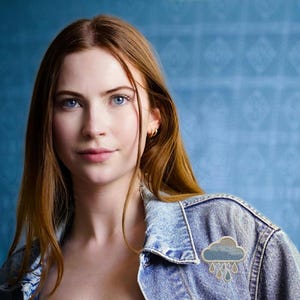 May include: A light blue denim jacket with a cloud and rain drop patch. The patch is embroidered with shades of blue and gray. The background is a textured blue.