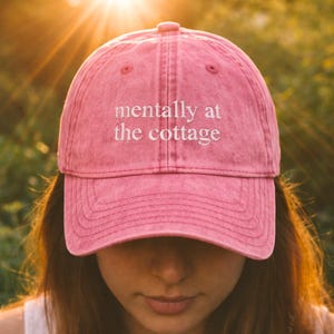 Könnte beinhalten: Rosa Baseballkappe mit dem weißen Schriftzug "mentally at the cottage". Die Kappe wird von einer Person mit langen braunen Haaren getragen, mit einem verschwommenen Hintergrund aus Grün und Sonnenlicht.