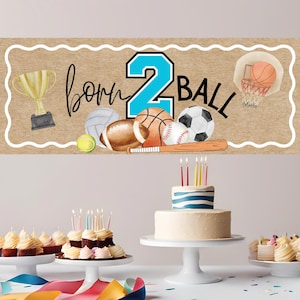 May include: A banner with a brown background and a white border features the text "born 2 ball" with sports-themed illustrations. A gold trophy, basketball, soccer ball, baseball, football, and tennis ball are depicted. A two-tiered cake with candles is in the foreground.