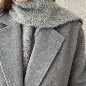 May include: A close-up of a gray wool coat with a matching gray textured scarf. The coat has a classic lapel design. The scarf is a lighter shade of gray and adds a cozy element to the outfit. The overall look is simple and elegant.