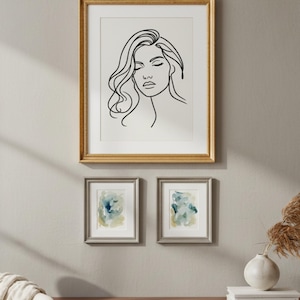 May include: Three framed art pieces on a wall. The largest is a gold-framed line drawing of a woman's face. Below are two smaller silver-framed abstract watercolor paintings in shades of blue and green. A modern home decor theme.