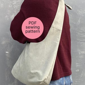 Peut inclure: Un sac en bandoulière gris clair avec une large sangle, porté sur l'épaule. Le sac est fait d'un matériau souple et a un design simple. L'image comprend un cercle rose avec le texte "PDF sewing pattern".