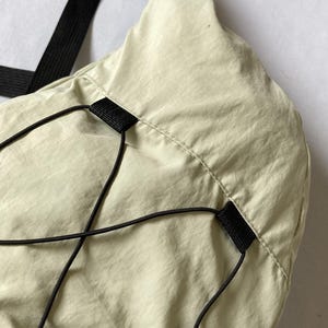 May include: Close-up of a light green shoulder bag with a black strap and drawstring closure. The bag is made of a smooth, lightweight material. Black drawstrings and a black strap are visible.