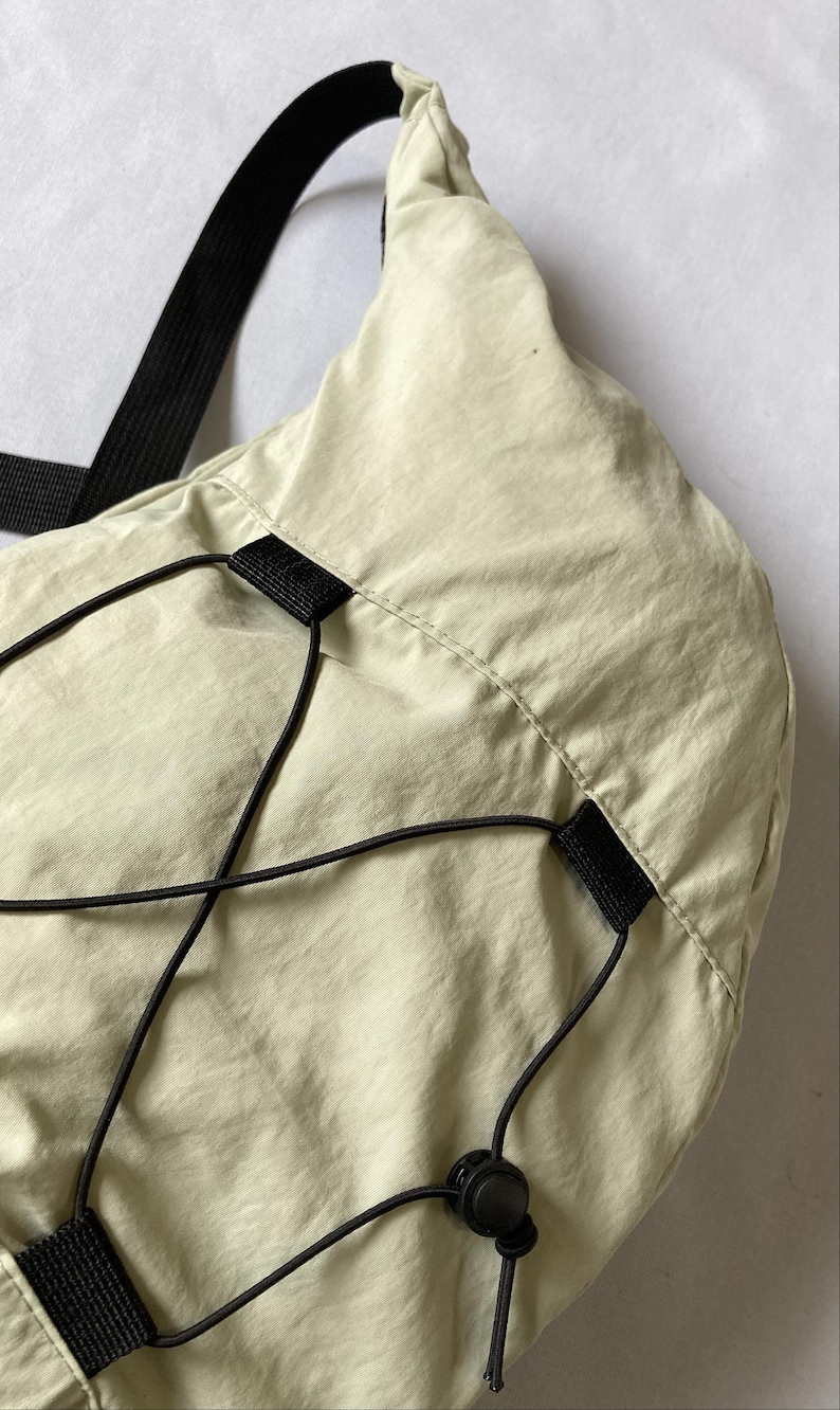 May include: Close-up of a light green shoulder bag with a black strap and drawstring closure. The bag is made of a smooth, lightweight material. Black drawstrings and a black strap are visible.