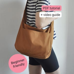 May include: A solid brown shoulder bag with a wide strap and an external pocket. The image includes text that reads PDF tutorial + video guide and Beginner friendly. The bag is worn over a striped t-shirt and black shorts against a plain background.