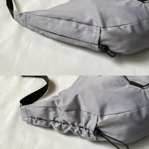 May include: A light gray crossbody bag with a black strap and drawstring closure. The bag is made of a smooth, possibly nylon, material. The bag has a rounded shape and a black drawstring closure.