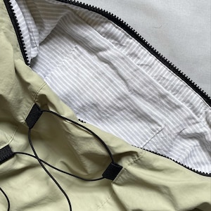 May include: A light green bag with a black zipper and drawstring closure. The interior lining is white with gray stripes. The bag has black straps and accents.
