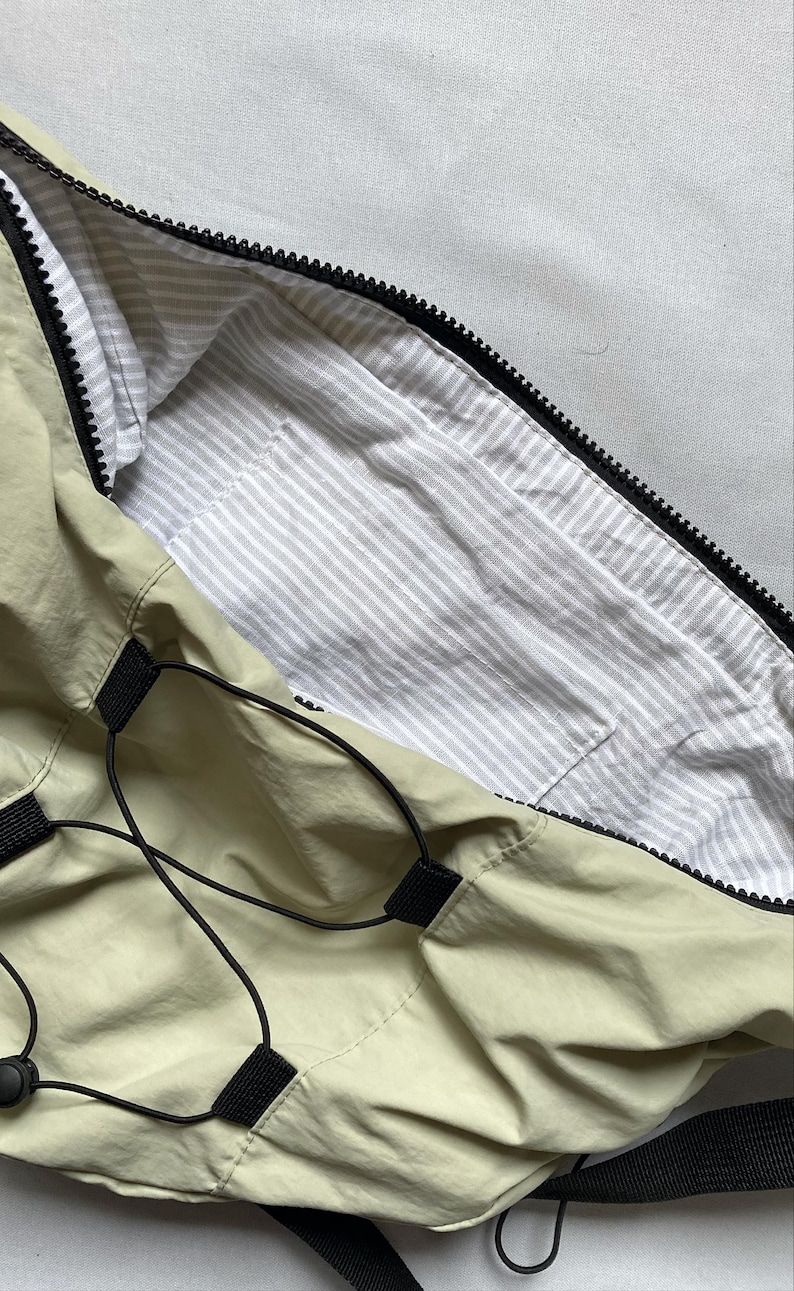 May include: A light green bag with a black zipper and drawstring closure. The interior lining is white with gray stripes. The bag has black straps and accents.