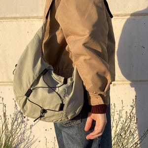 May include: A light green shoulder bag with a drawstring closure and black accents. The bag is worn over a tan jacket and blue jeans. The bag has a rounded shape and a black strap.
