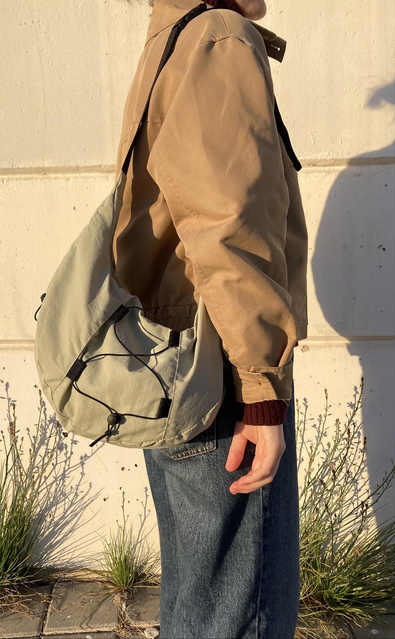 May include: A light green shoulder bag with a drawstring closure and black accents. The bag is worn over a tan jacket and blue jeans. The bag has a rounded shape and a black strap.