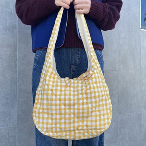 Pu&ograve; includere: Una borsa a tracolla trapuntata a quadretti gialli e bianchi. La borsa ha una forma arrotondata e un'ampia tracolla. La borsa &egrave; tenuta da una persona che indossa un gilet blu, un maglione bordeaux e jeans.