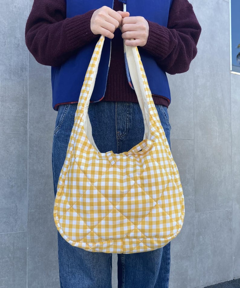 Pu&ograve; includere: Una borsa a tracolla trapuntata a quadretti gialli e bianchi. La borsa ha una forma arrotondata e un'ampia tracolla. La borsa &egrave; tenuta da una persona che indossa un gilet blu, un maglione bordeaux e jeans.