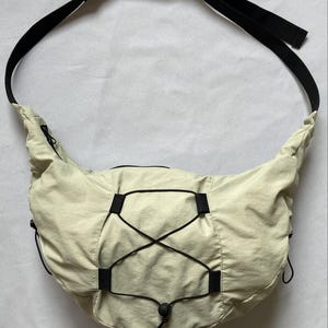 May include: A light beige crossbody bag with a black adjustable strap. The bag features a drawstring closure and black accents. The bag is made of a lightweight, possibly nylon, material.