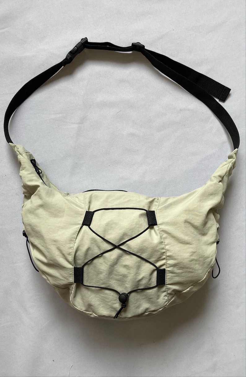 May include: A light beige crossbody bag with a black adjustable strap. The bag features a drawstring closure and black accents. The bag is made of a lightweight, possibly nylon, material.