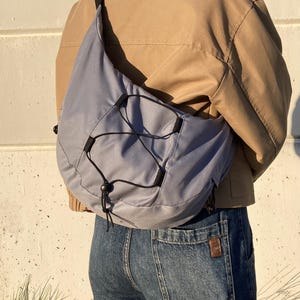 May include: A light blue crossbody bag with a black drawstring closure and black straps. The bag is worn over a tan jacket and blue jeans.