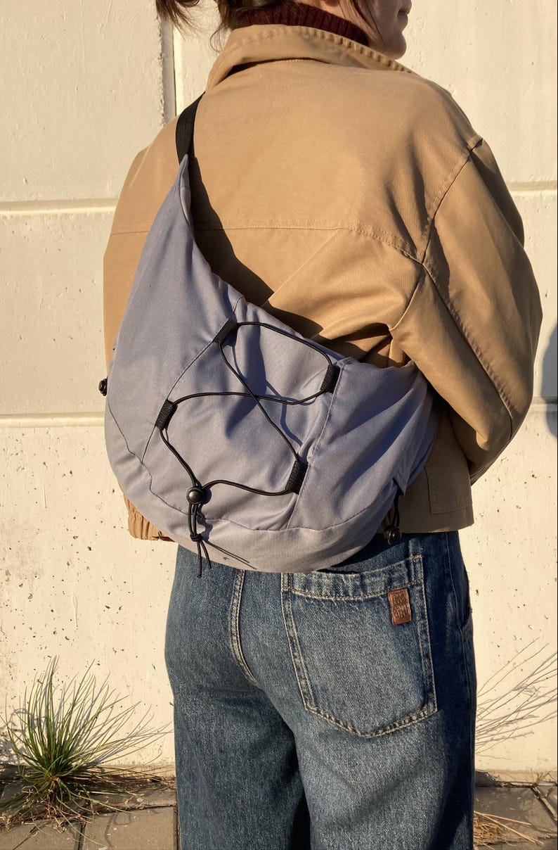 May include: A light blue crossbody bag with a black drawstring closure and black straps. The bag is worn over a tan jacket and blue jeans.