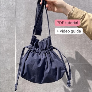 Puede incluir: Un bolso azul marino con cordón y correa larga, sostenido por una persona. El bolso tiene costuras blancas y una parte superior fruncida. El texto en la imagen dice "PDF tutorial + video guide".