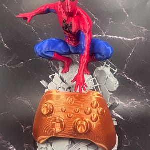 May include: A 3D printed figurine of a red and blue superhero in a crouching pose, positioned atop a gray rubble base. The figure is reaching down toward a metallic copper-colored video game controller that is integrated into the base structure.
